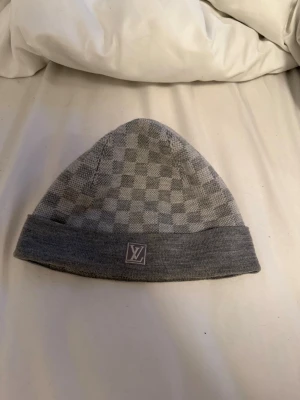 Grå Louis Vuitton mössa med rutmönster - Säljer en snygg grå mössa från Louis Vuitton med klassiskt rutmönster och vikt kant. Mössan har en diskret LV-logga framtill och är stickad i mjukt material som känns skönt mot huden. Perfekt accessoar för att hålla stilen när det är kallt ute.