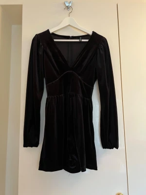 Svart sammetsjumpsuit med v-ringning - Supersnygg svart jumpsuit i mjuk sammet med lång ärm och djup v-ringning. Modellen har markerad midja och vida shorts, samt dragkedja i ryggen för enkel påklädning. Perfekt för dig som vill ha en trendig och bekväm look.