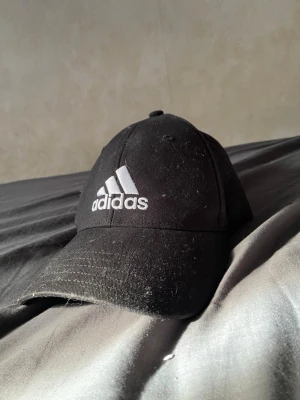 Svart Adidas keps med logga - Snygg svart keps från Adidas med klassisk böjd skärm och justerbar rem bak. Framtill sitter den ikoniska Adidas-loggan broderad i vitt. Tillverkad i bomullsmaterial som andas. Perfekt accessoar för en sportig och avslappnad stil.