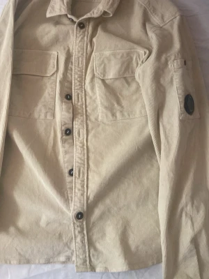 Beige overshirt från C.P. Company - Stilren beige overshirt i manchestertyg från C.P. Company. Två bröstfickor med lock, knappstängning framtill och ikonisk linsdetalj på ärmen. Perfekt lager-på-lager-plagg med avslappnad passform och klassisk krage.