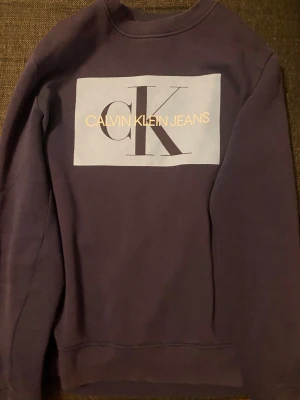 Blå sweatshirt från Calvin Klein - Snygg blå sweatshirt från Calvin Klein Jeans med stort vitt tryck och CK-logga på bröstet. Tröjan har rund halsringning, långa ärmar och ribbade muddar. Perfekt för en avslappnad och trendig look.