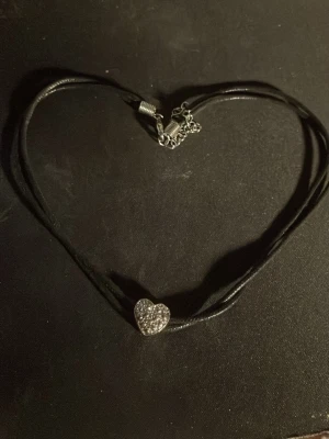 Halsband med hjärthänge och pärlor - Snyggt halsband med svart band och ett glittrande hjärthänge dekorerat med små pärlor. Halsbandet har justerbart spänne i silverfärg och passar perfekt för att ge din outfit lite extra bling.