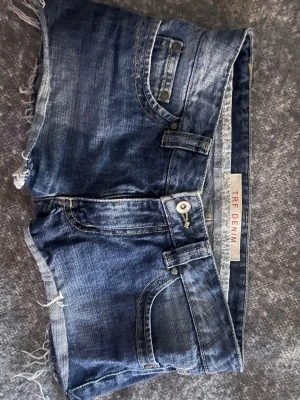 Blå jeansshorts från TRF Denim - Säljer ett par klassiska blå jeansshorts från TRF Denim med råa, fransiga benslut och slitna detaljer. Shortsen har fem fickor, bälteshällor och knappgylf. Perfekta till sommaren för en avslappnad look.