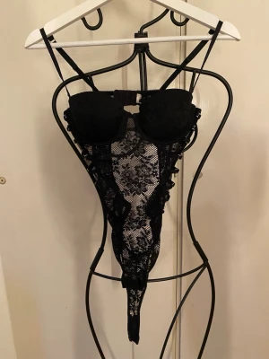 Svart spetsbody med banddetaljer och öppen rygg  - Säljer en svart body i spets med vadderade kupor och smala axelband. Bodyn har genombruten spets med blommönster, dekorativa band i sidorna och en stor rosett bak. Materialet är stretchigt och ger en snygg siluett.