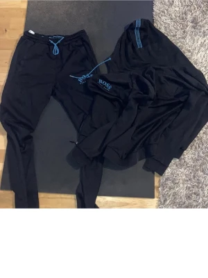 Svart loungewear-set från BOSS - Snyggt svart loungewear-set från BOSS med matchande byxor och tröja. Setet har blå snörning i midjan och blå logga på både byxa och tröja. Perfekt för chill dagar hemma eller när du vill vara bekväm men ändå se fräsch ut.