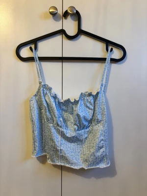 Blommig blå linnetopp med smala band - Söt blå linnetopp med små vita blommor och smala axelband. Toppen har en croppad passform och lätt rynkad kant upptill för en feminin look. Perfekt för varma dagar och enkel att styla med jeans eller kjol.