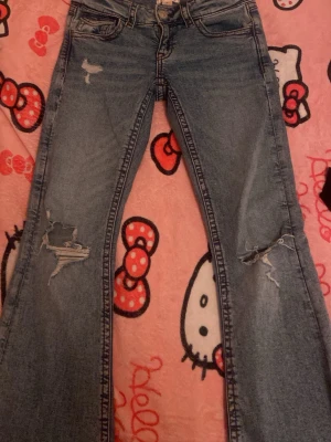 Blå slitna bootcut jeans Gina Tricot - Säljer ett par blå bootcut jeans från Gina Tricot med slitningar och hål på båda benen. Jeansen har klassisk femficksmodell, låg midja och snygg tvättad look. Perfekta för en avslappnad och trendig stil.