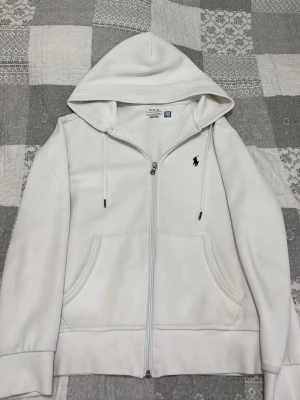 Vit hoodie från Polo Ralph Lauren - Snygg vit hoodie med dragkedja från Polo Ralph Lauren. Klassisk design med huva och dragsnören, två fickor framtill och den ikoniska lilla loggan broderad på bröstet. Perfekt för en clean och stilren look. Skriv om ni vill ha mer bilder😁Det finns en liten defekt vid arm mudden men syns knappt!!!