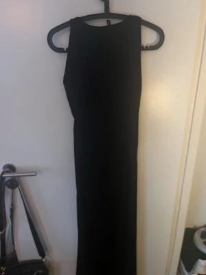 Svart långklänning med hög hals - En stilren svart långklänning med hög hals och ärmlös design. Klänningen har en rak och elegant siluett som ger en klassisk look. Perfekt för dig som gillar minimalistisk stil och vill ha en enkel men snygg klänning.