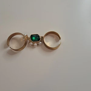 Guldfärgad dubbelring med grön sten och pärlor - En elegant guldfärgad dubbelring med en rektangulär grön sten i mitten och små vita pärlor som dekoration. Ringen är designad för att bäras på två fingrar samtidigt.