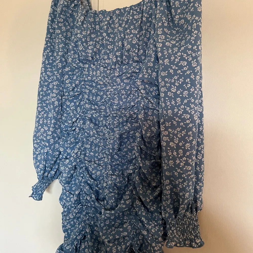 En blå klänning med vita små blommor på. Sitter smickrande på kroppen med hjälp av stretchen och modellen. Kan användas både som på bild men ocksp off shoulder om man drar ner armarna. Aldrig använd och väldigt fint skick! . Mekot.