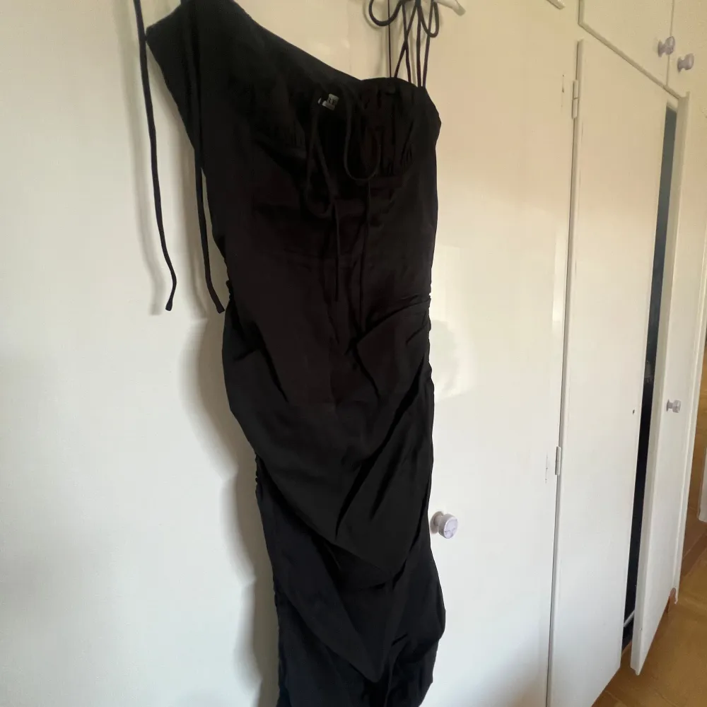 En svart adoore klänning med tunna band som knyts över axeln. Dragkedja i sidan. Längden går att justera genom knyten i sidan. ALDRIG ANVÄND - lappar sitter på!!! Pris går att diskutera. . Mekot.