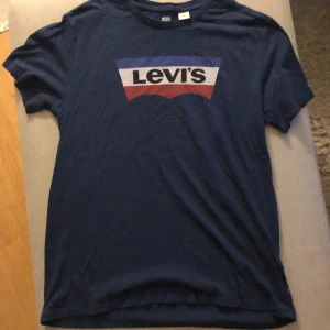 Mörkblå t-shirt från Levi's - En mörkblå t-shirt från Levi's med korta ärmar och en tryckt logotyp i vitt, rött och blått på framsidan. T-shirten är tillverkad i Turkiet.