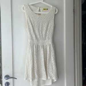 Vit spetsklänning - En coquette vit spetsklänning med ärmlös design och rund halsringning. Klänningen har en öppen rygg med knappar och en kort längd. Perfekt för sommar och fest tillfällen.
