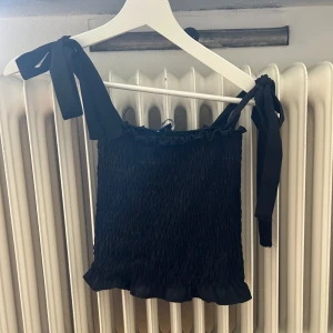 Svart tubtopp med knytband - En svart tubtopp med knytband på axlarna. Toppen har en rynkad design och volangdetaljer vid nederkanten.