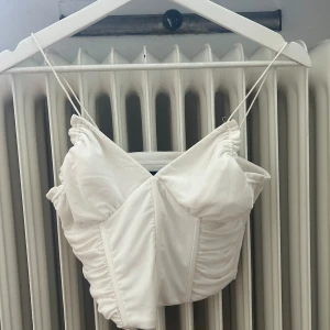 Vit korsett - En elegant vit korsett med smala axelband och rynkade detaljer. Korsetten har en tight passform och är perfekt för festliga tillfällen.