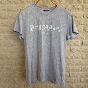 Grå t-shirt från Balmain - En stilren grå t-shirt från Balmain med vit text 'BALMAIN PARIS' tryckt på framsidan. T-shirten har korta ärmar och en rund halsringning.