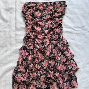 Blommig klänning från Blind Date Casual - En blommig klänning från Blind Date Casual med rosa och svarta blommor. Klänningen har volanger och är axelbandslös.