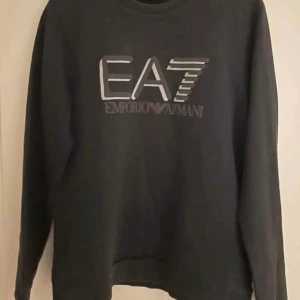 Svart tröja från Emporio Armani - En svart långärmad tröja från Emporio Armani med logotypen 'EA7' tryckt på framsidan. Tröjan har en rund hals och är tillverkad i ett bekvämt material.
