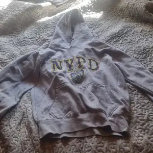 En bekväm grå hoodie från NYPD med en stor ficka framtill och en huva. Tröjan har texten 'NYPD' och en logotyp tryckt på framsidan.