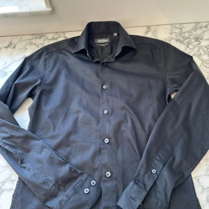 En svart skjorta från dressman  - En nattsvart skjorta från dressman i storlek S. Skjortan är mjuk och lätt att röra sig i. Den funkar perfekt för finare tillfällen respektive fester. Skjorta har jag endast använt 3-4 gånger så den funkar som spillrans ny. Passformen är tajt och den 