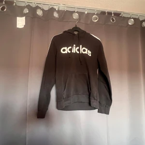 Svart hoodie från Adidas - En svart hoodie från Adidas med vit text på framsidan. Hoodien har en justerbar huva med dragsko och en stor ficka på magen. Den har långa ärmar och ribbstickade muddar vid ärmslut och nederkant.