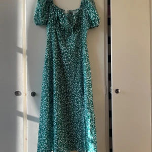 Grön blommig klänning från Shein - En grön långklänning från Shein med ett vitt blommönster. Klänningen har korta ärmar och en knytning framtill vid halsen. Den är tillverkad i ett lätt och bekvämt material, perfekt för sommarens varma dagar. Inte använd! 