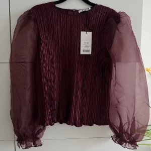 Topp från NAKD - Topp i ”vinröd” färg med lätt genomskinliga armar från NA-KD. Helt oanvänd och i storlek 36.