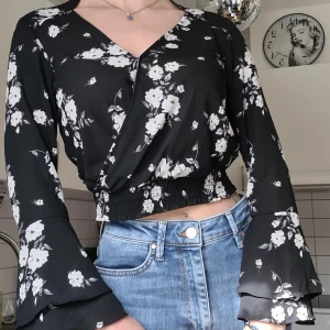 Blommig blus - Vacker blus med vita blommor och svart bakgrund. Utsvängda ärmar, stängs vid bröstet med en liten knapp. Går in i midjan, kortare modell. Stolek: XS-S, använd men i bra skick. Kom med prisförslag 😊  Material: 100% Polyester. 