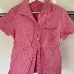 En kortärmad rosa skjorta i frotté från Nelly.com (Terry Beach Set) med knappar framtill samt en bröstficka och en krage. Använt 1 gång, som ny. Nypris för hela setet: 599 kronor.