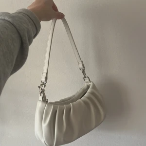 Vit handväska - Vit handväska med 39 cm lång axelrem som är avtagbar. Köpt utomlands för ett par år för 200kr. Mått är 26x14 cm.  Inga innerfickor. Väskan går att stänga med en dragkedja. Materialet är fejkläder. Använd få gånger. Skickar fler bilder vid intresse❤️💞