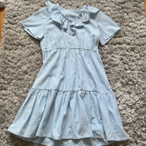 Vit, blå randig volang klänning - Jag säljer min fina volangklänning på grund av att jag beställt fel storlek, har testat den 1 gång men tyvärr vart den för liten. Storlek 140 och pris kan diskuteras.