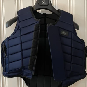Marinblå ridväst - Säljer nu min marinblåa ridväst då den inte kommer till användning. Köptes på en tävling 2022, använd 1 gång sen dess. Väldigt fint skick utan några skavanker.  Storlek 12, lite osäker på vad de innebär i vanlig storlek, men passar mig som har 32/34