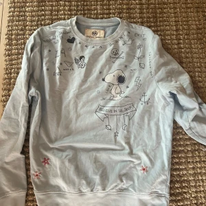 Ljusblå tröja från Taylor Tweed med tryck - En ljusblå tröja från Taylor Tweed med tryck av olika symboler och text, inklusive en tecknad figur och texten 'Believe in yourself'. Tröjan har långa ärmar och rund halsringning.