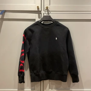 Svart Ralph lauren med röd text på sidan - Ralph lauren med röd text på armen där det står ”polo”. fint skick, knappt använd. Köpt på NK nypris 1299