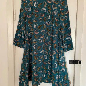 Söt klänning från H&M - Söt klänning med kameleonter från H&M Stl 36 (S) men lös passform över midja och höfter. Knapp i nacken och fint veckad bak samt fina detaljer framtill. Använd ett fåtal gånger, i nyskick Grundfärgen är blågrön, typ petrol. Längd: 92cm