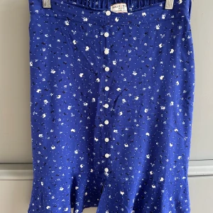 Blå Blommig kjol med knappar  - Nyskick, aldrig använd. Blå kjol med vita blommor och små svarta detaljer från Holly&whyte . Kjolen har vita knappar framtill och en slits nere. 
