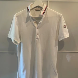 Moncler Piké - Riktigt fin Moncler piké i färgen vit. Otroligt snyggt med ränderna vid kragen. Tröjan är i nyskick utan några defekter alls. Var inte rädd att skriva ifall ni har några frågor. Storlek M✅