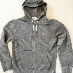 Maison Kitsune zip hoodie  - Jag säljer min gråa maison kitsune hoodie. Skicket är otroligt bra utom en liten vit fläck på vänstra framsida (bild 2). Jag köpte den på Paul and friends för 1800. Mitt pris är 400.