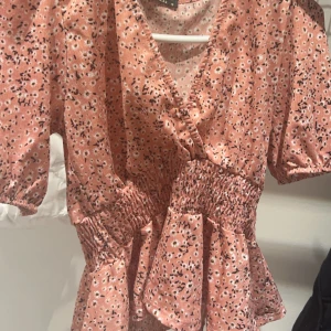 Rosa blommig topp - En rosa blommig topp med korta ärmar och v-ringning. Toppen har en smockdetalj i midjan och volang nedtill.