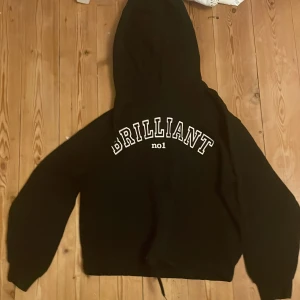 Svart hoodie från Lindex - En svart hoodie från Lindex med texten 'BRILLIANT no1' tryckt på framsidan. Tröjan har långa ärmar, en luva och en rosett längst ner.