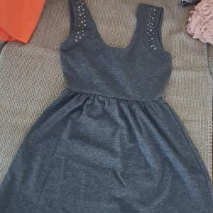 Mörkgrå klänning med nitar - En mörkgrå klänning med nitar på axelbanden. Klänningen har en rund halsringning och är ärmlös. Den är av normal passform och har en elastisk midja.