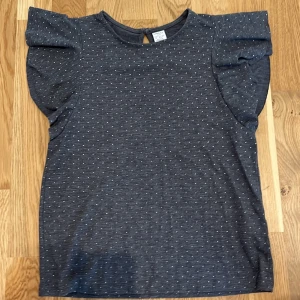 Grå prickig topp från Lindex - En grå topp från Lindex med små vita prickar. Toppen har korta ärmar med volangdetaljer och en knappstängning i nacken.