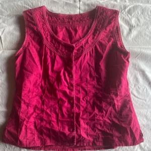 Röd ärmlös blus - En ärmlös blus med knappar framtill och dekorativa sömmar runt halsringningen och ärmhålen. Blusen har en v-ringad hals och är tillverkad i ett lätt och bekvämt material.