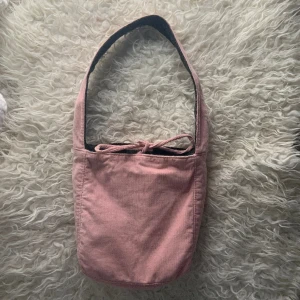 Söt rosa väska  - Söt rosa axelremsväska. Den har snören som kan knytas samt en ficka med dragkedja i. Bra skick förutom pytte liten defekt som visas på bild 2. Syns knappt. 