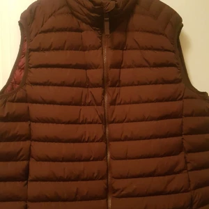 Brun och grå dunväst från Dressmann och Kappahl - Två dunvästar, en brun från Dressmann och en grå från Kappahl. Båda västarna har dragkedja och är quiltade. Den bruna västen har en röd insida medan den grå västen har svarta detaljer på fickorna och axlarna.