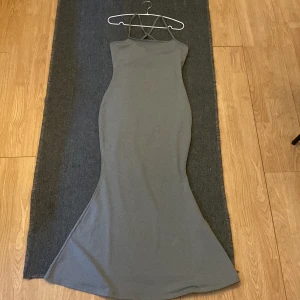 Grå långklänning med korsade axelband - En elegant grå långklänning med korsade axelband. Klänningen har en figurnära passform och är ärmlös, skims