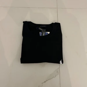 Svart t-shirt från Polo Ralph Lauren - En klassisk svart t-shirt från Polo Ralph Lauren med en liten broderad logotyp på vänster bröst. T-shirten har en rund hals och korta ärmar.