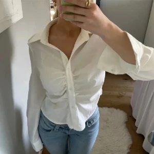 Vit blus med knappar och rynkdetalj - Säljer en snygg vit figursydd blus med knappar och en cool rynkdetalj framtill. Blusen har långa ärmar och en v-ringad krage. Perfekt för både vardag och fest! Passar bra till jeans eller kjol. 💕Lånade bilder!