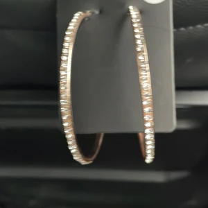 Stora guldiga hoop-örhängen med stenar - Säljer ett par stora, guldiga hoop-örhängen med glittrande stenar längs hela framsidan. De är perfekta för att piffa upp vilken outfit som helst och passar både till vardags och fest. Örhängena är i nyskick och har en elegant och stilren design.